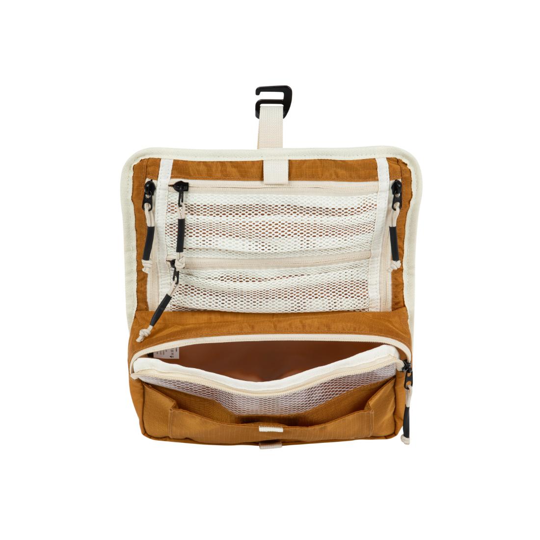 תיק רחצה לנסיעות מסוג "Vagabag" של Aztec בצבעי חום בהיר ולבן, עם כיסים ותאים מרשת בעלי רוכסן, מוצג על רקע לבן. התיק תלוי על וו שחור בחלקו העליון.