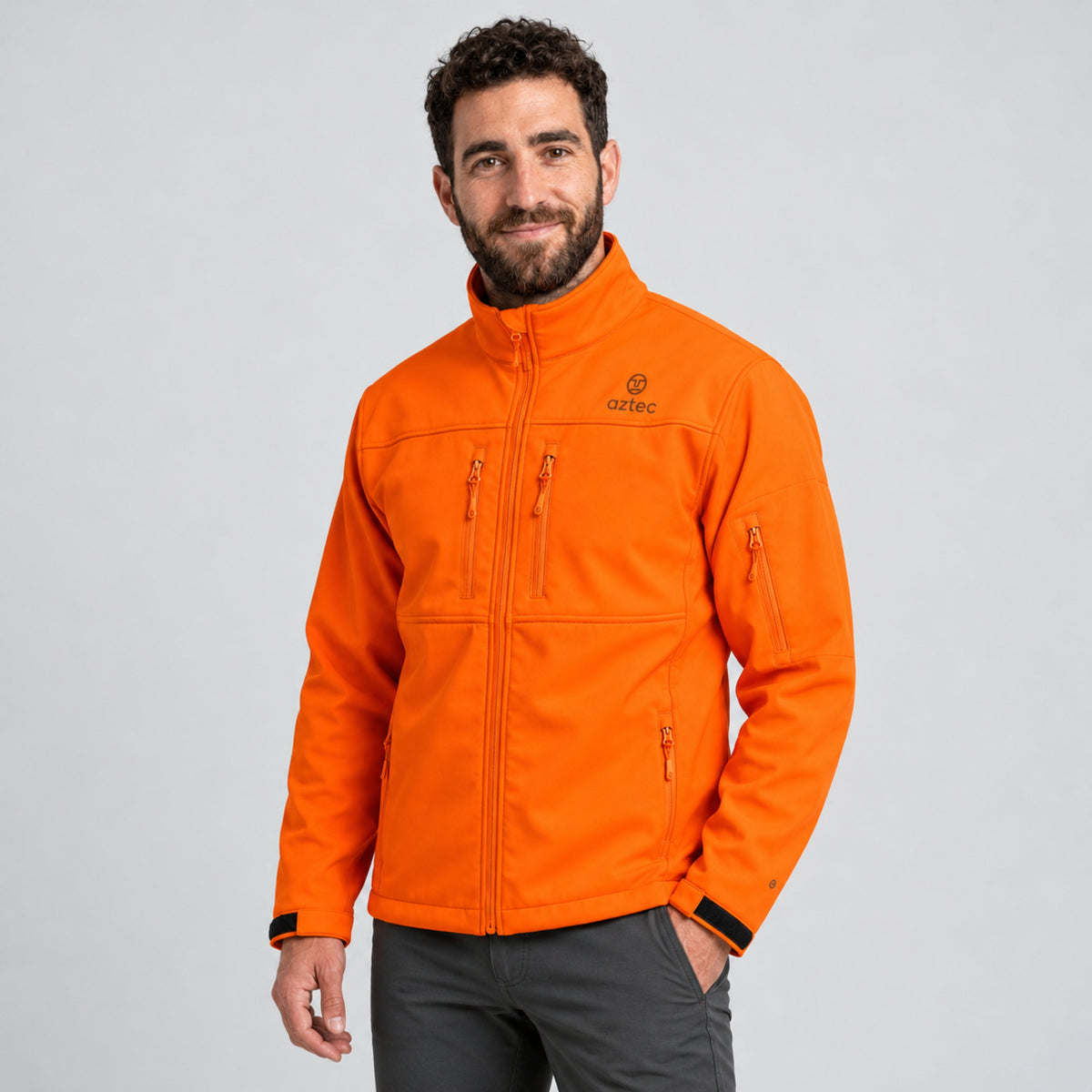 גבר לבוש במעיל Aztec Recovery Softshell בצבע כתום בוהק, עם כיסים בחזה ובשרוולים, בשילוב עם מכנסיים אפורים.