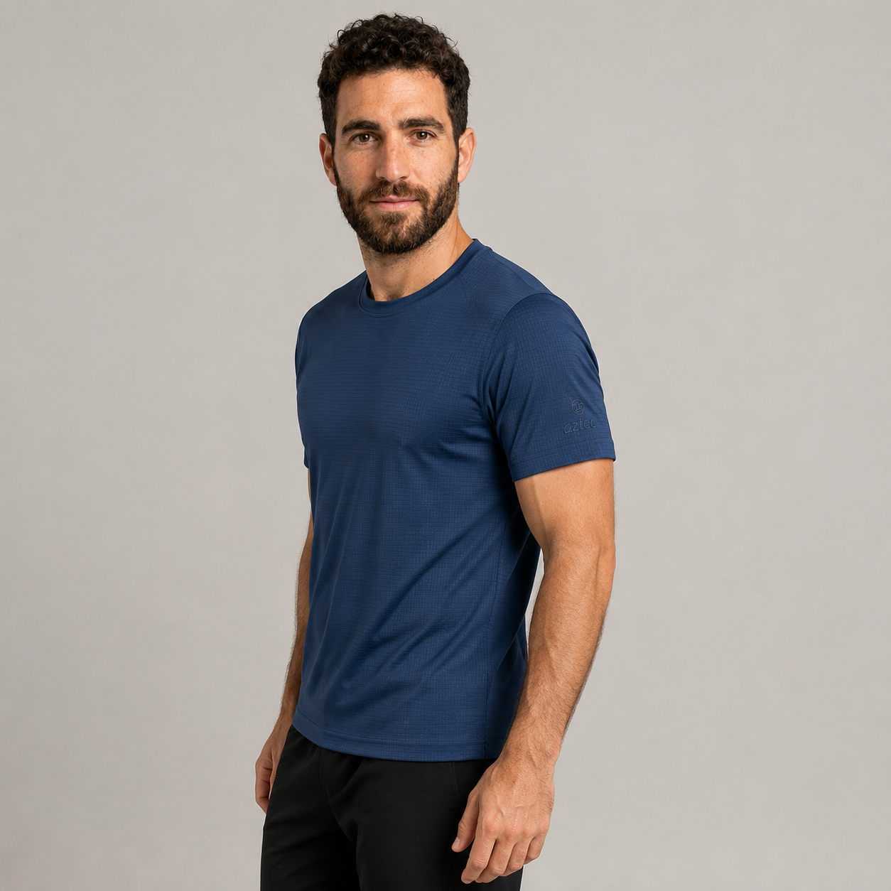 גבר עם שיער קצר ומתולתל וזקן לובש את החולצה הכחולה “Aztec Flow M” ומכנסיים שחורים, עומד על רקע אפור בהיר אחיד ומביט במצלמה בחיוך קל.
