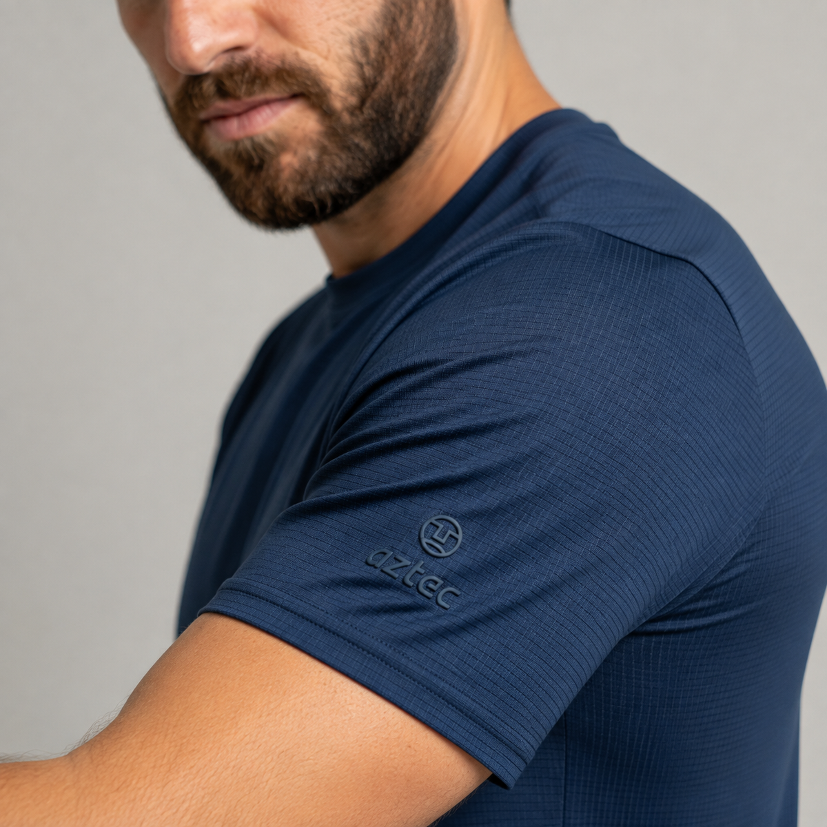 גבר מזוקן לובש חולצת ספורט צמודה עם שרוולים קצרים מדגם “Flow M” בצבע כחול-צי של המותג Aztec, כאשר לוגו המותג והכיתוב “Aztec” נראים על שרוולו. הרקע הוא אפור בהיר אחיד.
