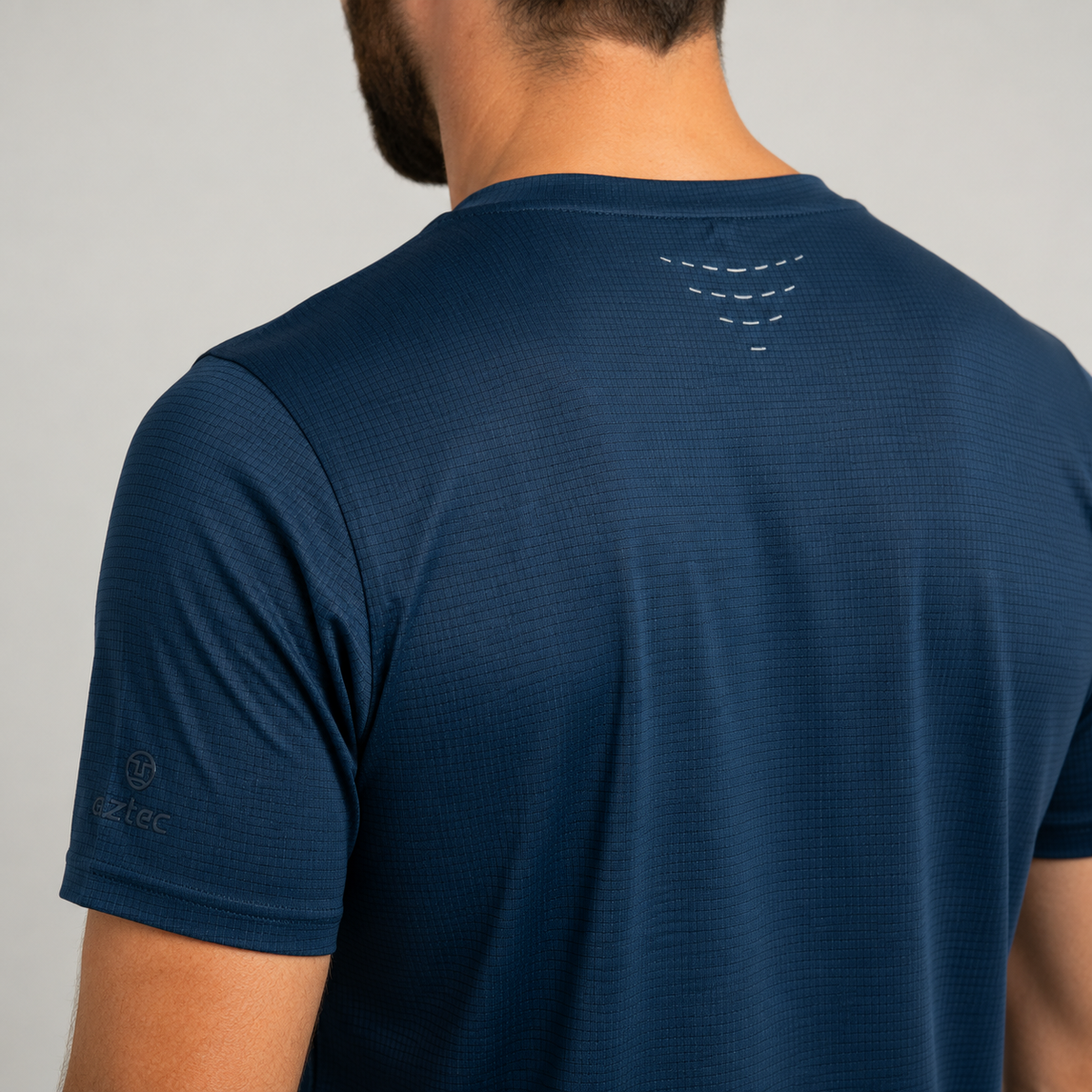 בתמונה נראה גבר מאחור, לבוש בחולצת הספורט Aztec Flow M – חולצה ספורטיבית בצבע כחול כהה עם שרוולים קצרים, הכוללת עיצוב מחזיר אור קטן בקרבת הצוואר ולוגו עדין על השרוול.