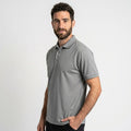 גבר בעל שיער קצר וכהה וזקן, לבוש בחולצת Aztec Darco Polo II S/S M עם שרוולים קצרים בצבע אפור ובמכנסיים שחורים, עומד על רקע לבן חלק, כשידו האחת בכיסו.