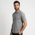 גבר בעל שיער קצר וכהה וזקן, לבוש בחולצת Aztec Darco Polo II S/S M עם שרוולים קצרים בצבע אפור ובמכנסיים שחורים, עומד על רקע לבן חלק, כשידו האחת בכיסו.