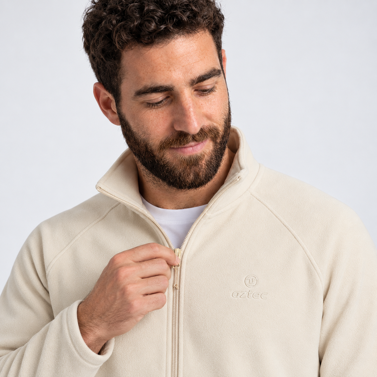 גבר עם שיער מתולתל וזקן רוכסן את מעיל הפליס “Aztec Raven Full Zip” בצבע שמנת, המעוטר ברקמה בסגנון אצטקי, כשהוא מביט מטה ומחייך קלות על רקע בהיר ופשוט.