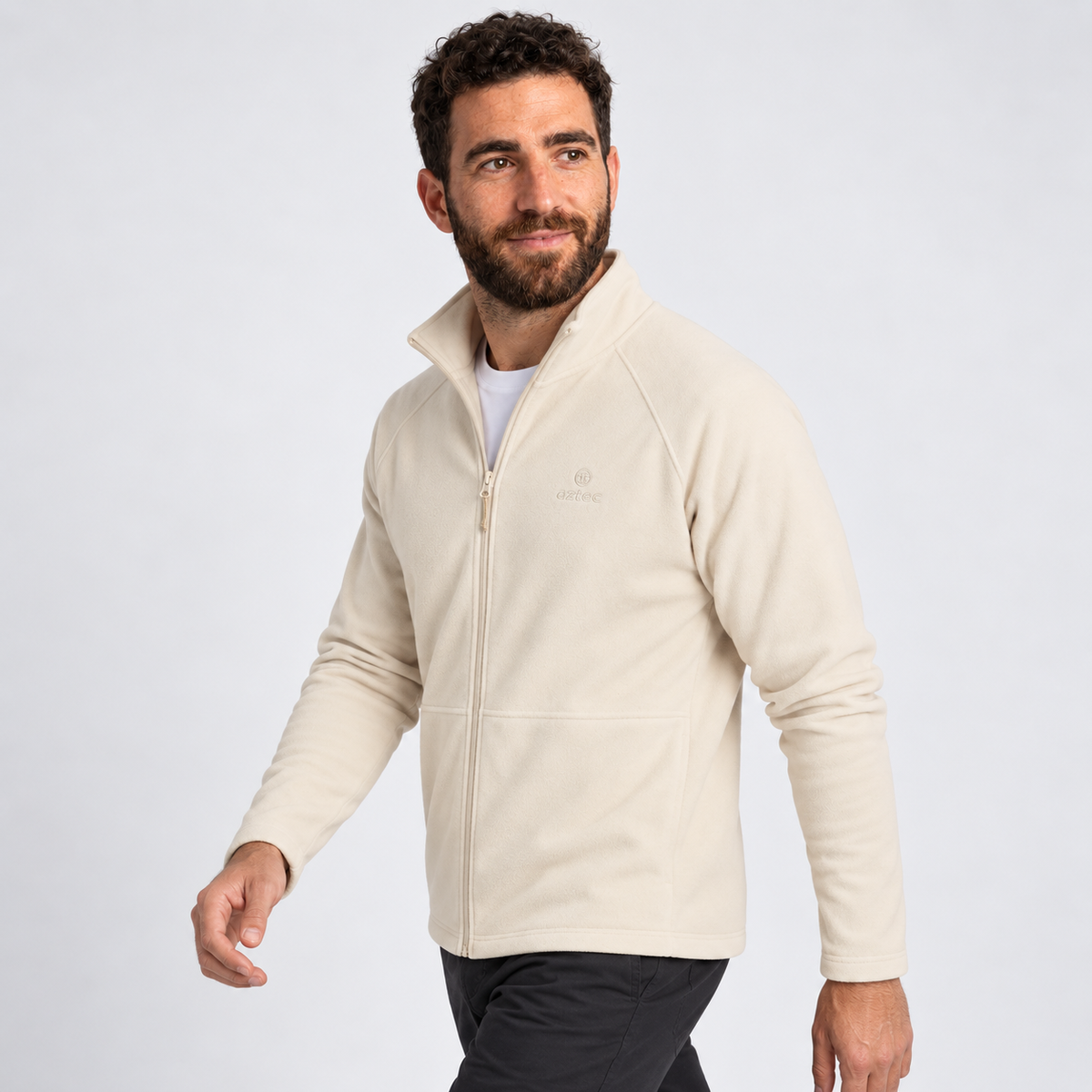 גבר עם שיער קצר ומתולתל וזקן לובש מעיל פליס “Aztec Raven Full Zip” בצבע בז' בהיר ומכנסיים שחורים, הולך ומביט לשמאלו על רקע אפור בהיר אחיד.