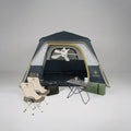 מתקן קמפינג הכולל את אוהל Aztec Bundle ATLAS VENT 4P בצבעים כחול ואפור, בליווי שני כיסאות מתקפלים בצבע בז', שולחן קטן עם ספלים וכלי מטבח, מחצלת ירוקה, תיק שחור ומאוורר תקרה בתוך האוהל — הכול מסודר על רקע אחיד.